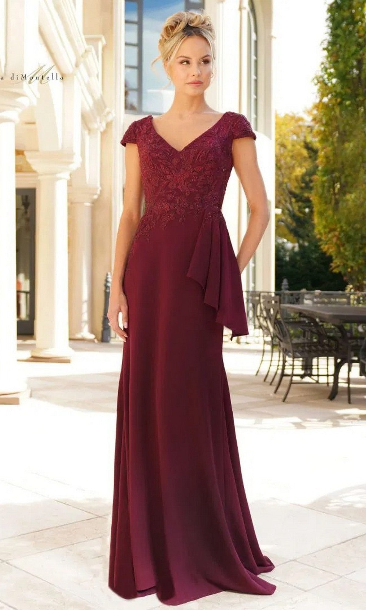 A woman stands outdoors in the Rina Di Montella RD2960 long formal dress in wine, featuring a stretch crepe skirt, lace cap sleeves, and an embroidered bodice. Her blonde hair is elegantly styled in an updo.